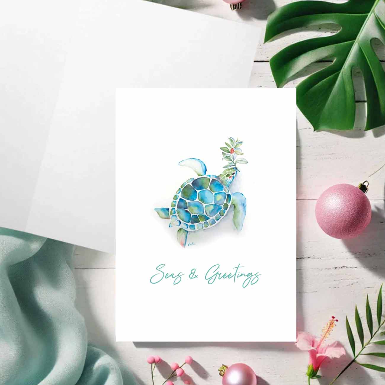 Flat lay of a beach Christmas cards featuring a watercolor sea turtle in shades of turquoise blue over a crisp white background. The words Seas & Greetings are set in trendy script type. Art and design by Victoria Grigaliunas of Do Tell A Bell a woman owned small business in Palm Beach Gardens, Florida.