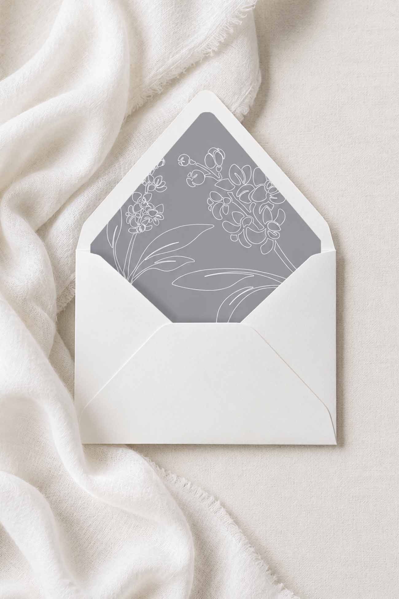 A white envelope with a gray floral envelope liner for wedding stationery, styled on a soft white fabric background.