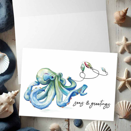 Flat lay of nautical Christmas cards designed with a watercolor blue octopus and holiday lights by artist Victoria Grigaliunas of Do Tell A Belle a woman owned small business in Southern Florida.