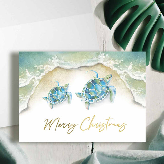 Flat lay of watercolor sea turtle Christmas cards with a beach shoreline in teal blue designed with artwork by Victoria Grigaliunas of Do Tell A Belle a woman owned small business in Palm Beach, Florida