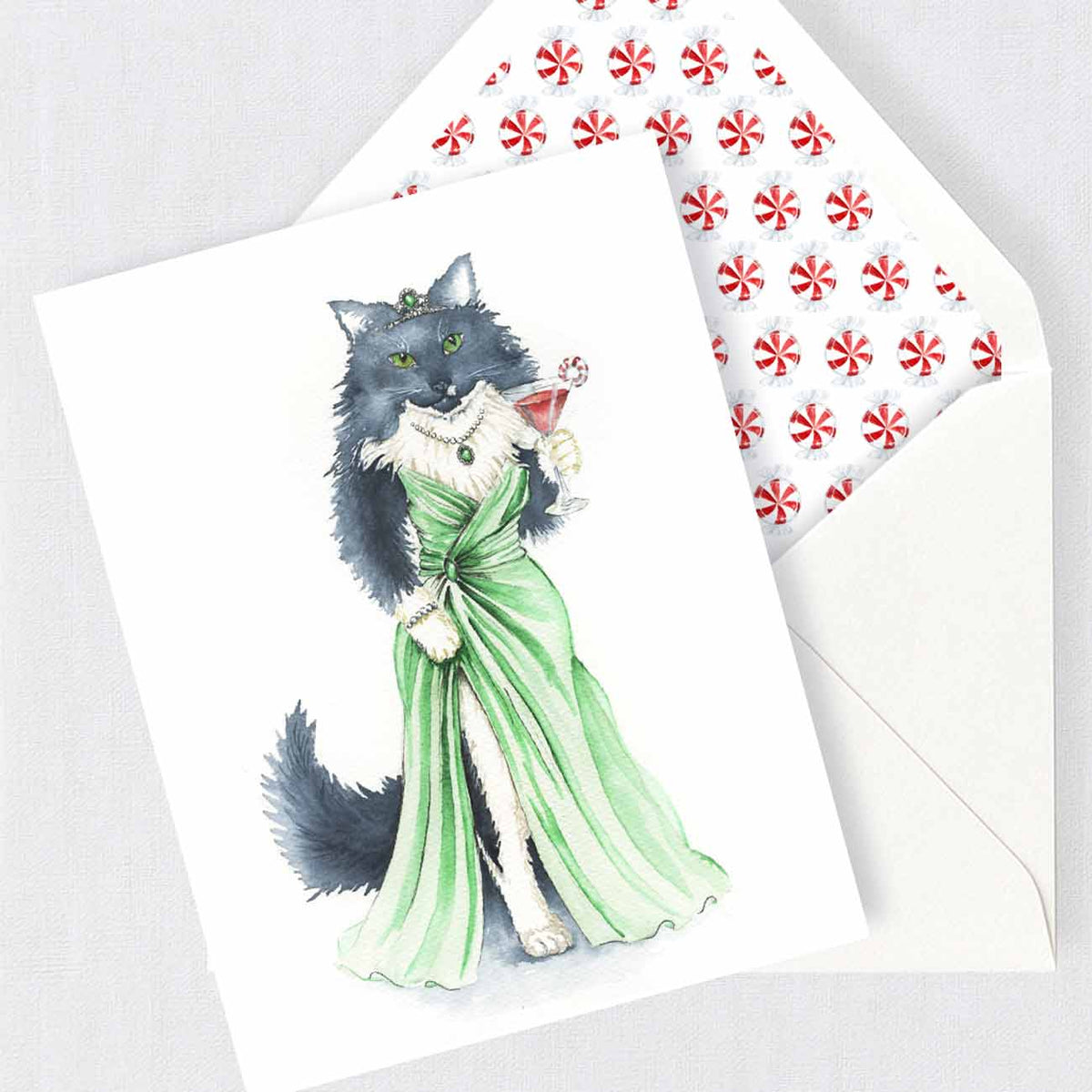 A white folded cat Christmas card featuring a watercolor illustration of a black cat in a green dress holding a martini glass, shown with a matching white envelope lined with a red-and-white candy cane pattern.