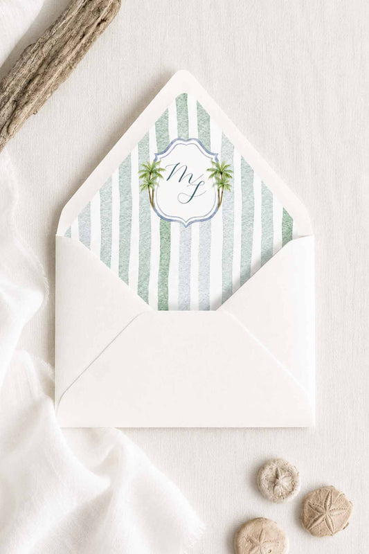A white envelope and envelope liner with stripes and watercolor monogram with in script between two palm trees, styled on a light fabric surface with draped white cloth, sand, and small natural beads.