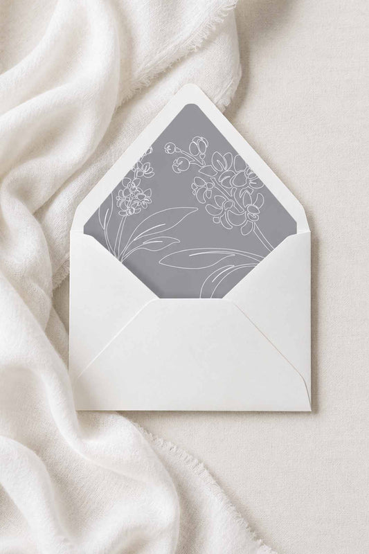 A white envelope with a gray floral envelope liner for wedding stationery, styled on a soft white fabric background.