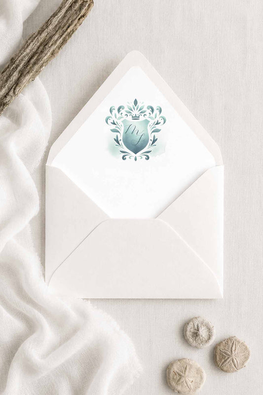A white envelope with a decorative teal green monogrammed crest printed on the front, styled on a light fabric surface with draped white cloth, driftwood, and small sand dollars.