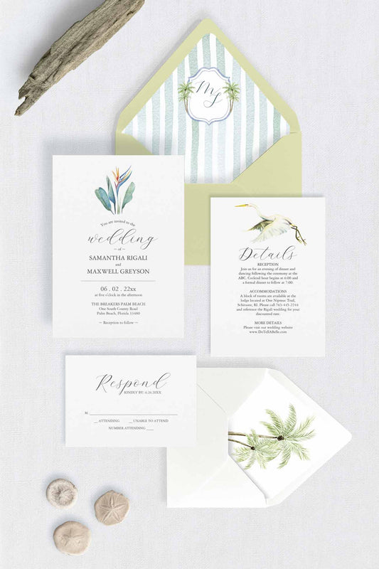 A coordinated wedding invitation suite laid flat on a light neutral surface, including a watercolor wedding invitation card with tropical floral artwork, a details card with a watercolor great white heron, a response card, a striped and monogram envelope liner with palm trees and pale green envelope, and a small rsvp card with white envelope and palm tree envelope liner. 