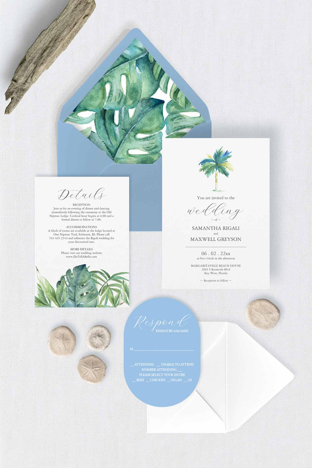 A tropical-themed wedding stationery set arranged on a light surface, including a light blue envelope with green monstera leaves, a white invitation card with a palm tree and script text, a wedding details card with green botanical artwork, a light blue oval rsvp card, and small natural stones and driftwood props.