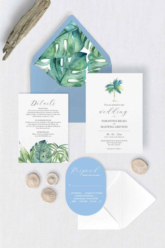A tropical-themed wedding stationery set arranged on a light surface, including a light blue envelope with green monstera leaves, a white invitation card with a palm tree and script text, a wedding details card with green botanical artwork, a light blue oval rsvp card, and small natural stones and driftwood props.