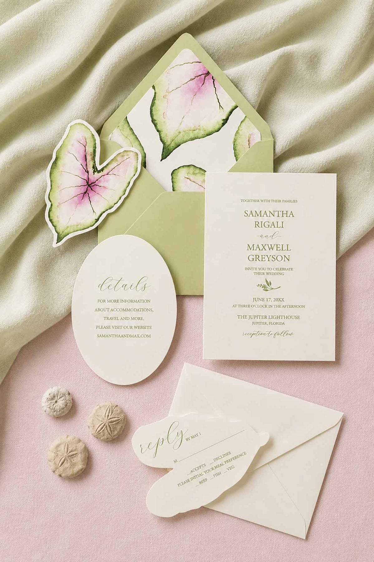A styled flat-lay of a botanical pink and green wedding invitation suite printed on cream cardstock with green envelopes and caladium leaves envelope liner and laser cut rsvp cards. The set includes a invitation card, an oval details card, a caladium leaf-shaped RSVP card, and a green envelope.