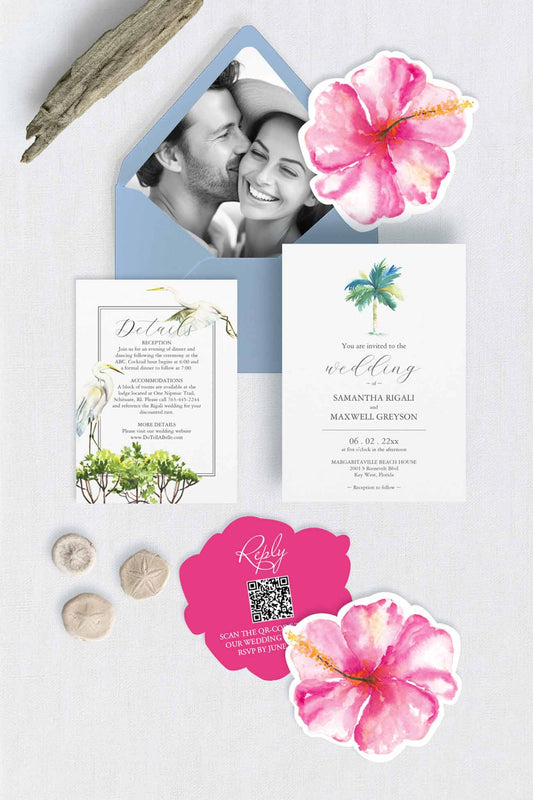 Tropical-themed wedding invitation suite with pink hibiscus flowers die cut rsvp card with qr code, palm tree wedding invitations, details card with tropical birds and Florida mangroves and and picture envelope liner with blue envelope on a pale grey background, styled with driftwood and sand dollars.