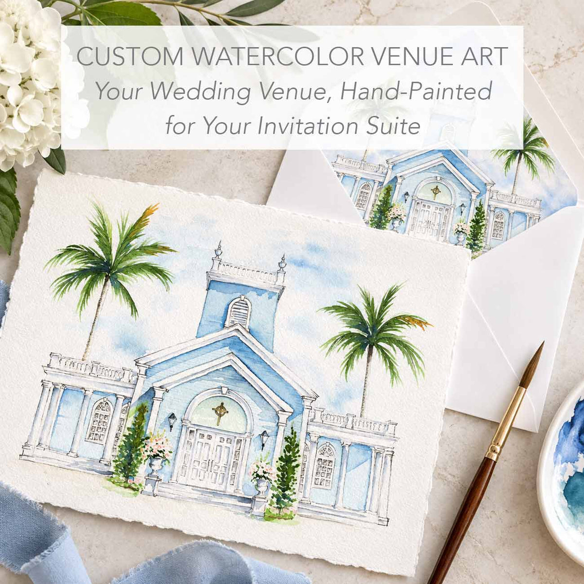 A watercolor art print of a blue church with palm trees, styled on a marble surface with envelope, envelope liners, a paintbrush, white flowers, and a text overlay reading 'CUSTOM WATERCOLOR VENUE ART'.